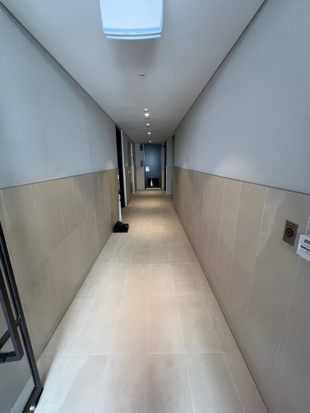 From preceding door facing straight. Broom obstructing ahead. Button to exit on right wall. Bathroom door on right wall at end of hallway ahead.