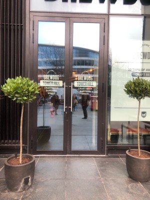 Glass doors with dark grey frame. Pull metal handle in centre to open. Plants to left and right.