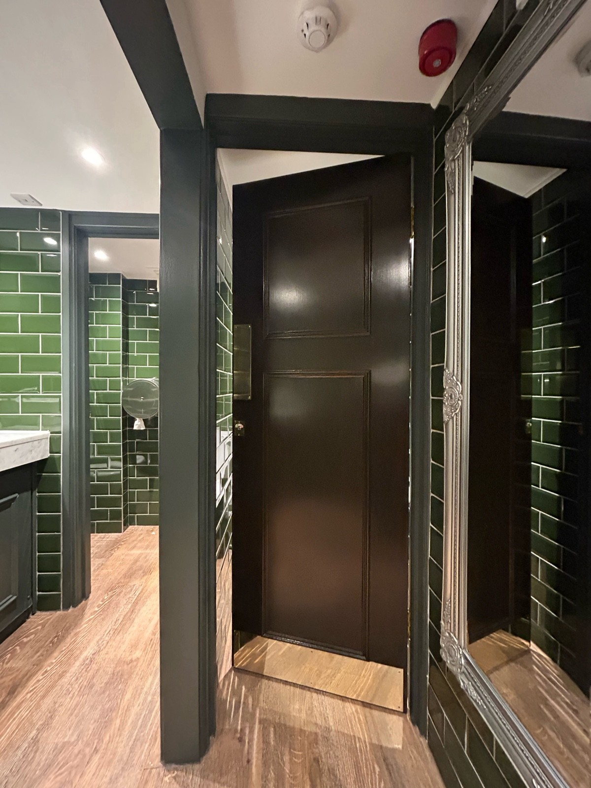 From preceding door facing right. Toilet cubicles ahead and on back left. Dark wooden door ahead, push from left to open. Sinks on left.