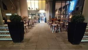 From street facing venue. Planters and fence on left and right. Tables and chairs behind on both sides. Path ahead to entrance at back.