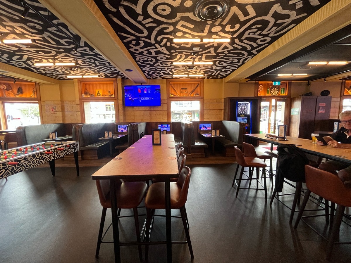 From after bar stools facing left. High tables with backed stools ahead to left and right. Booths with TVs on wall behind.