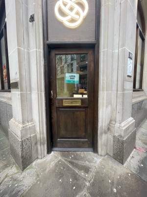 Facing single wooden door with glass panel. Push metal handle on left to open.