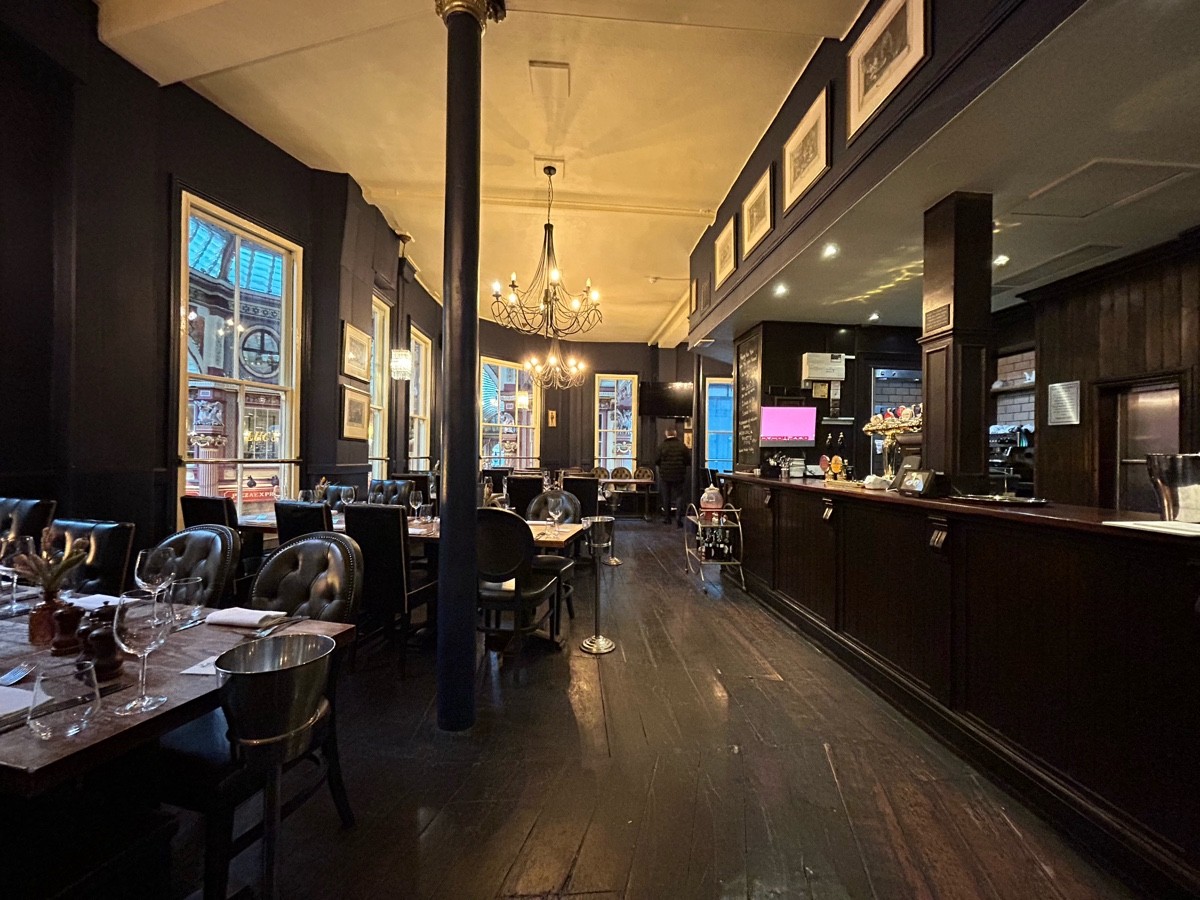 From doorway facing right. Bar on right. Two rows of tables and chairs on left, black pillar behind. Tables further ahead.
