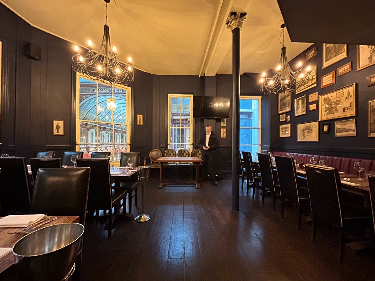 From end of bar facing straight. Tables and chairs along left and at back. Half booths on right. Black pillar to their left.