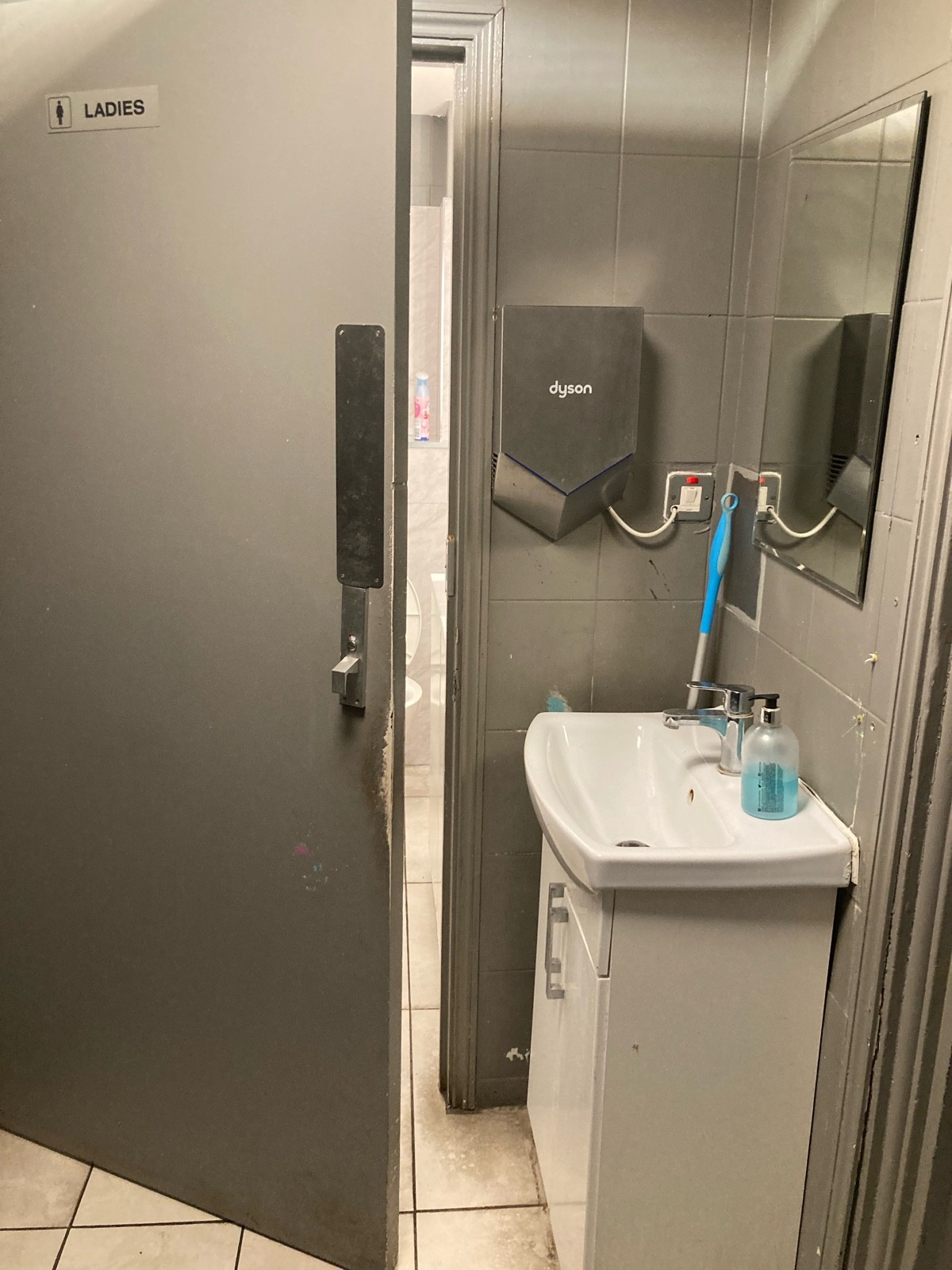 From door facing straight. Sink on right. Male’s door on left. Grey door ahead. Push door to open from right.