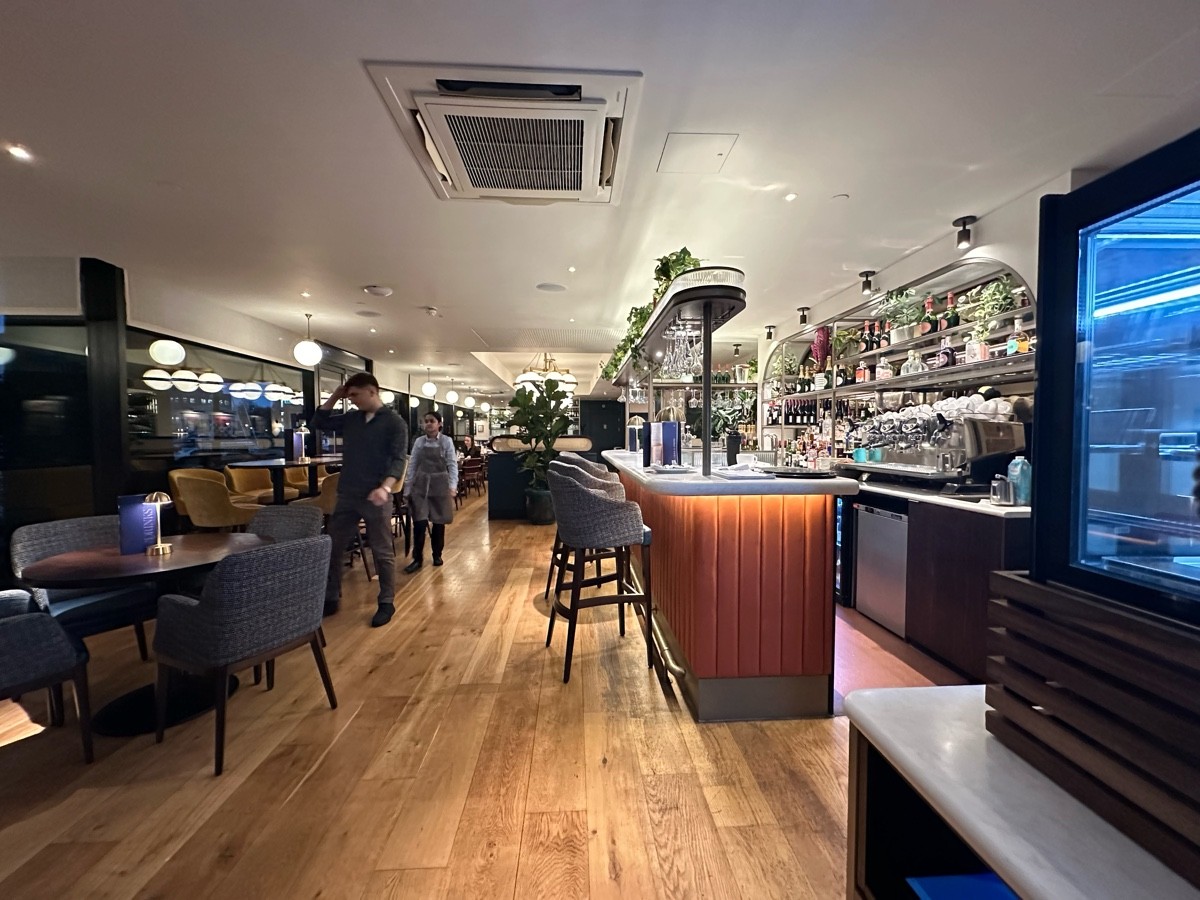 From archway facing straight. Bar with high stools along right. Tables and chairs along left.