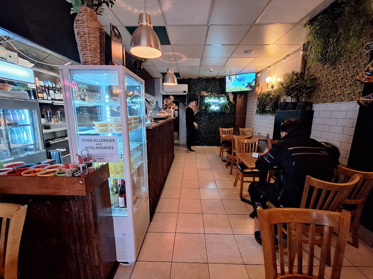 From toilet door facing straight. Counter and open kitchen on left. Tables and chairs along right.
