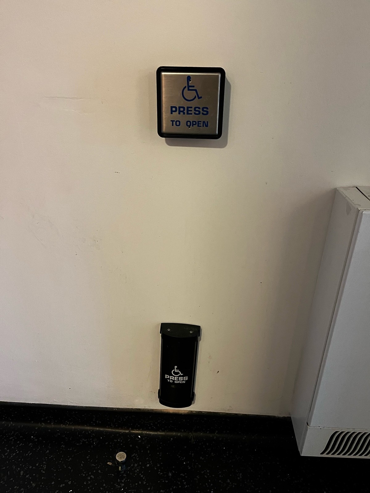 Two “Press to Open” buttons located next to bathroom door entrance. Top button is large, square and metal. Bottom button is black and near the floor.