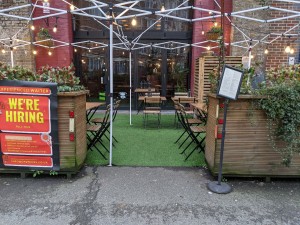 From street facing venue. Opening ahead with planters on both sides. Menu stand right of entrance.