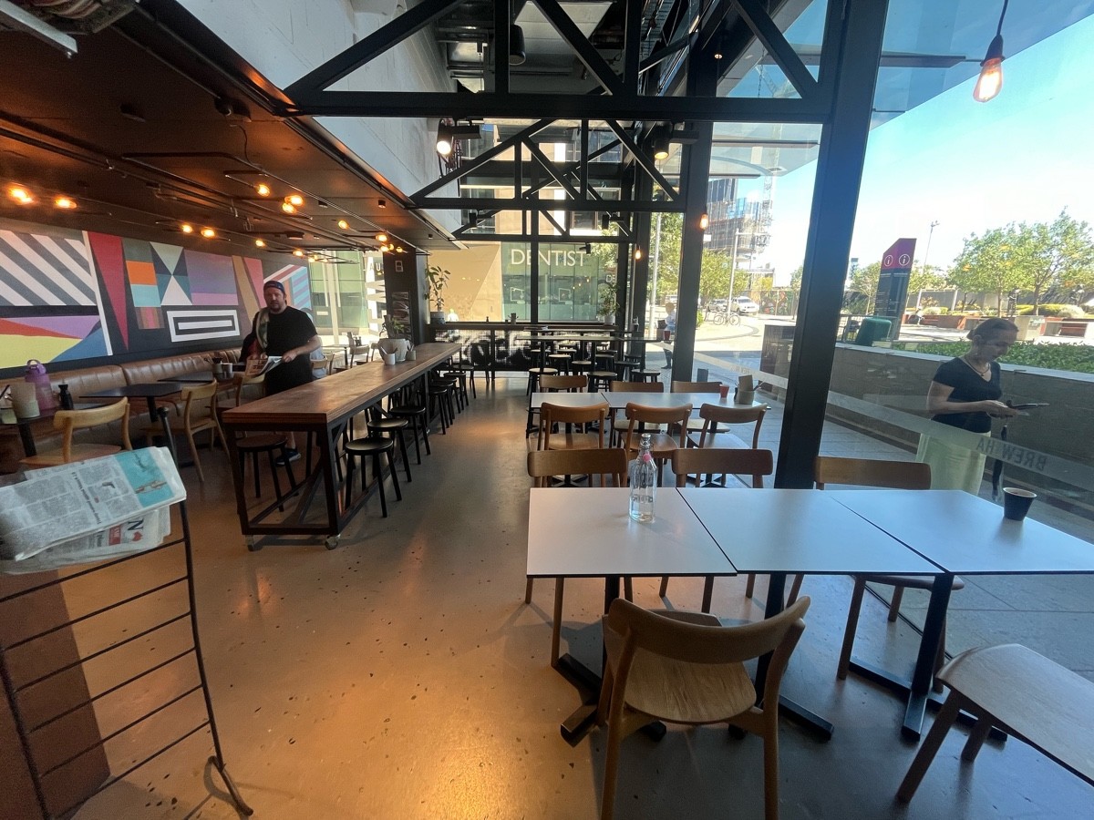 From after bins facing straight. Newspaper stand on left before tables and stools. Tables and chairs along right. High tables and stools on back wall