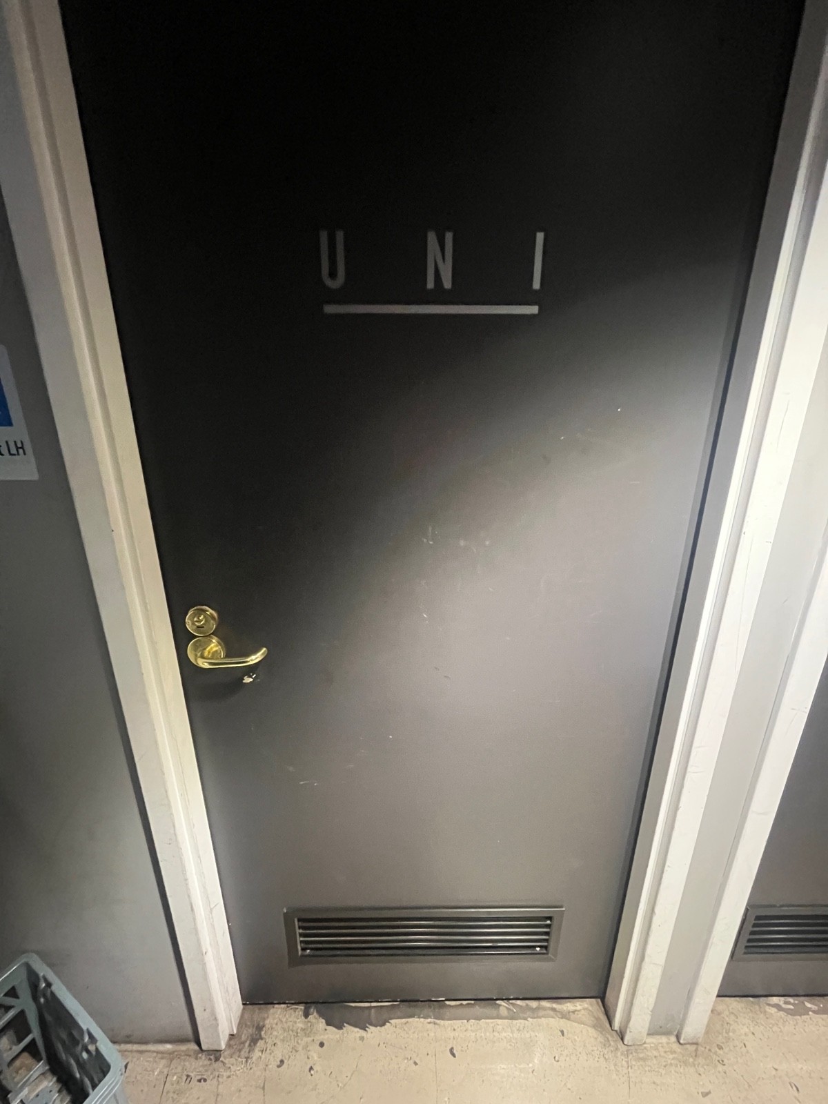 From end of hallway facing right. Grey wooden bathroom door with white frame and gold handle on left. Push to open.