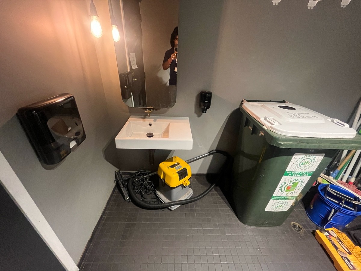 From door facing left. Sink on wall ahead. Vacuum cleaner under sink. High hooks on wall ahead to right obstructed by bins.