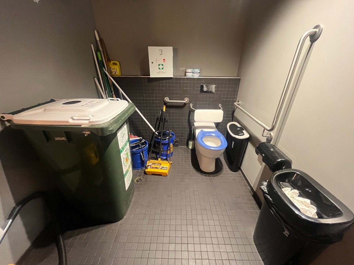 From door facing straight. Toilet on wall ahead. Bins and cleaning supplies obstructing transfer space.