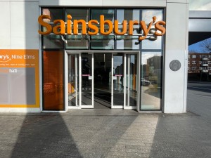 From street facing Sainsbury’s. White metal-framed glass doors ahead.
