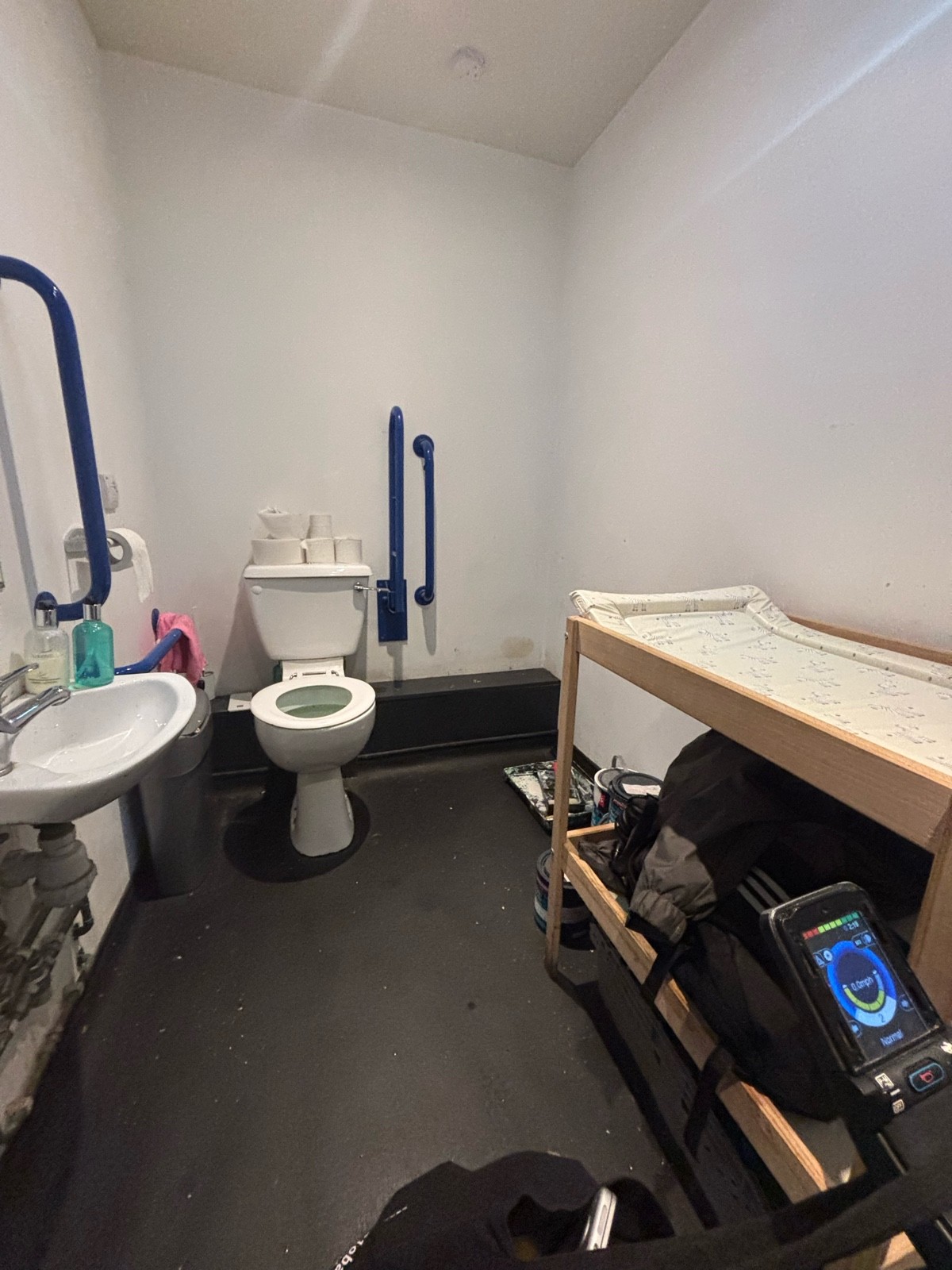 A wheelchair accessible toilet cluttered either objects that make it very inaccessible for a wheelchair user. Objects include paint cans & a table.
