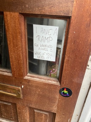 A sign on the door with a rainbow disability sticker and a sign saying “I have a ramp for wheeled access. Wave if you need it!”