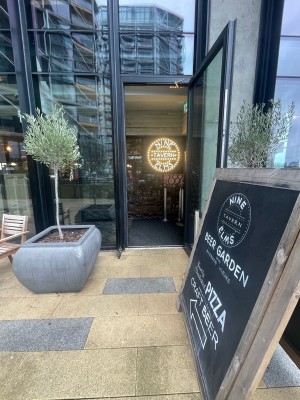 Glass door with black frame ahead. Pull silver metal handle on left to open. Planter on left before door. A-frame sign on right before door.
