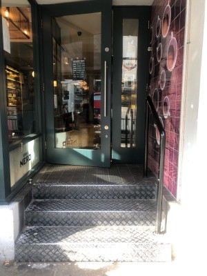 Three steps up to entrance. Black handrail along right. Glass door with dark green frame followed by doormat. Pull handle on right to open.