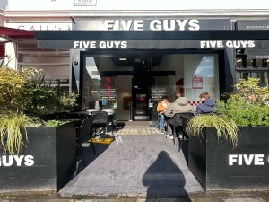 Black wooden planters on left and right. Tables and chairs beyond on both sides. Opening in centre. Entrance at back.