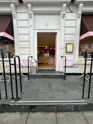 From pavement facing venue. Opening ahead. One step up ahead. Black railings to left and right.