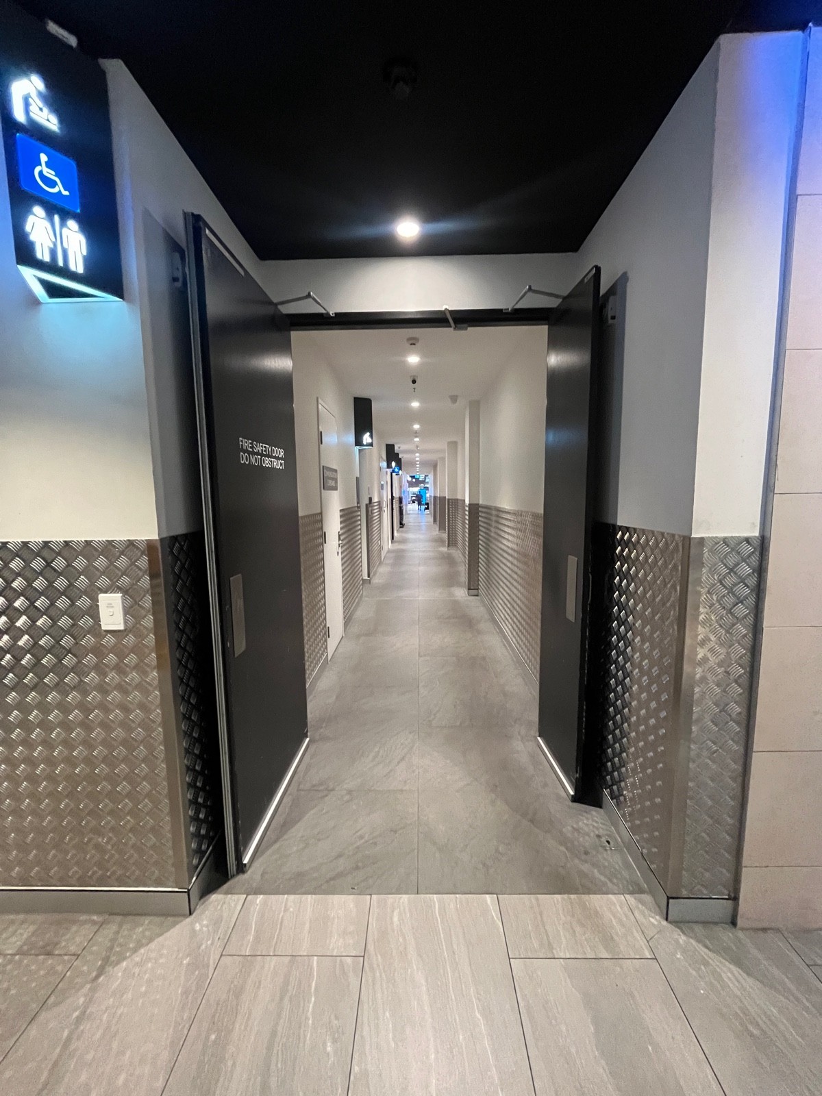 From after doors facing right. Second doors ahead. Grey wooden doors. Pull from middle. Doors typically open. Men’s bathroom on left wall ahead