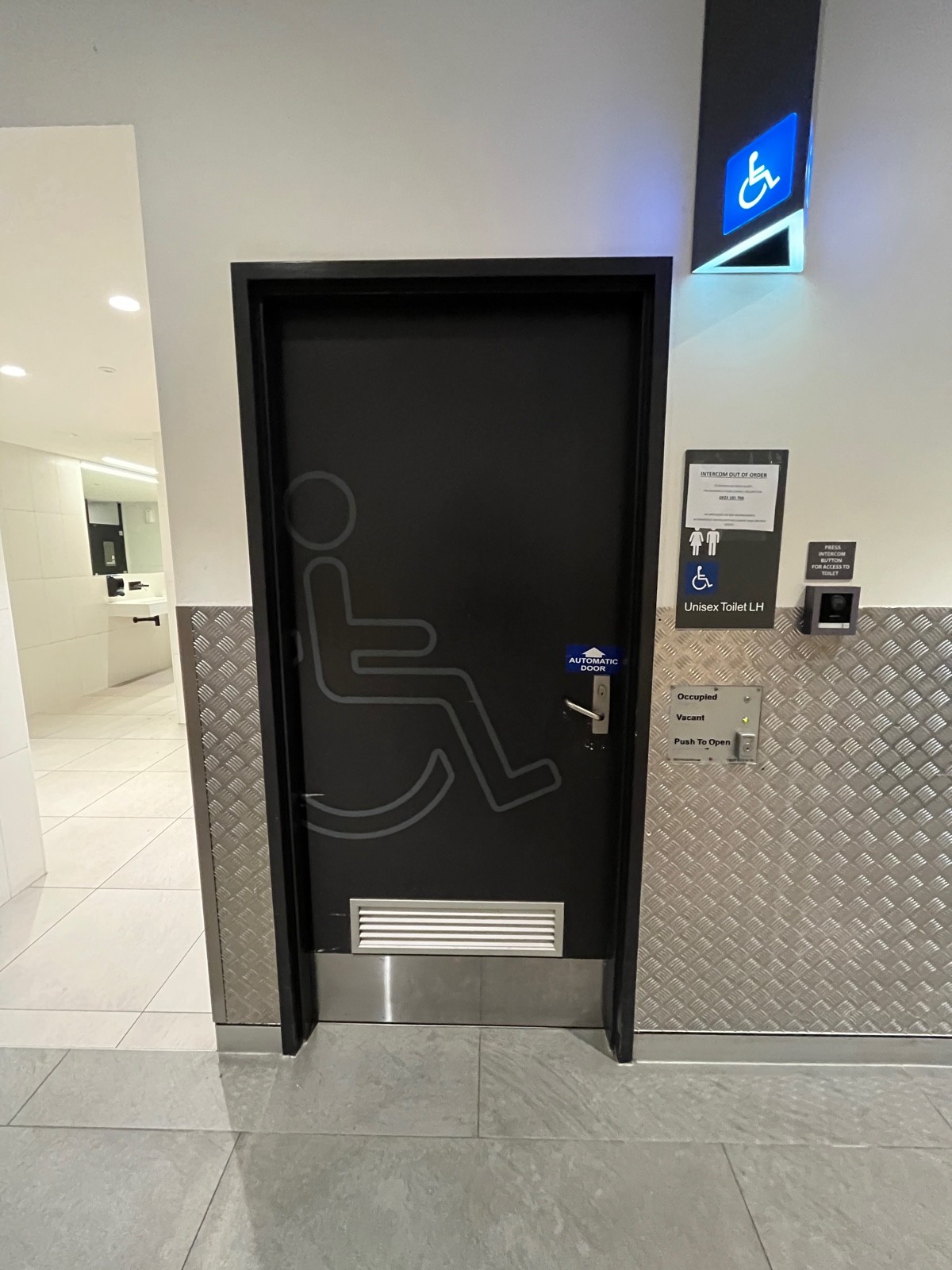 From after men’s bathroom facing left. Grey wooden bathroom door opens inwards from right. Intercom button for access on wall to right.