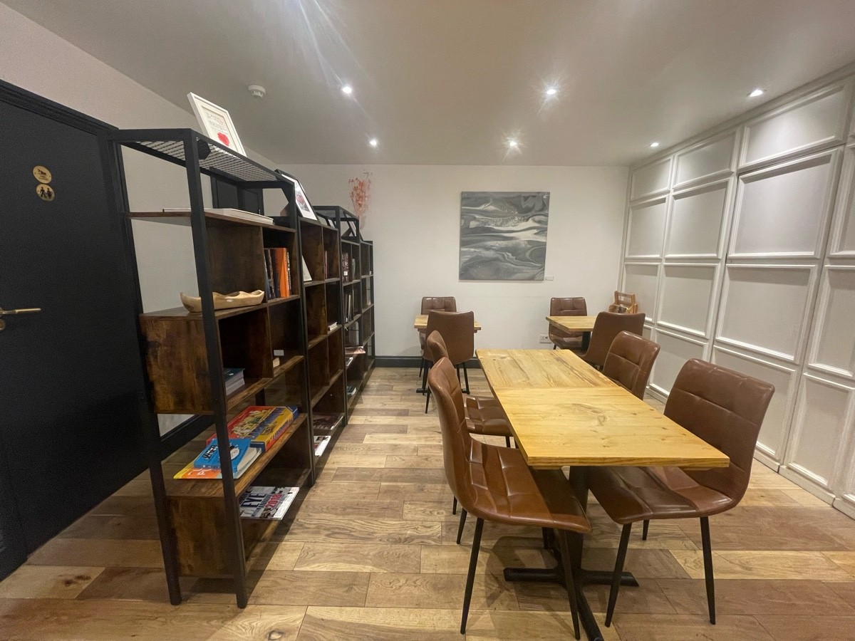 From after book shelf facing left. Tables and chairs ahead and along back. Metal book shelf to its left. Bathroom along left wall.