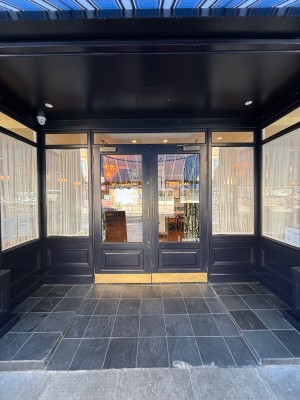 From after A-frame signs facing straight. A slope up to glass doors with black frames. Pull handles in centre to open.