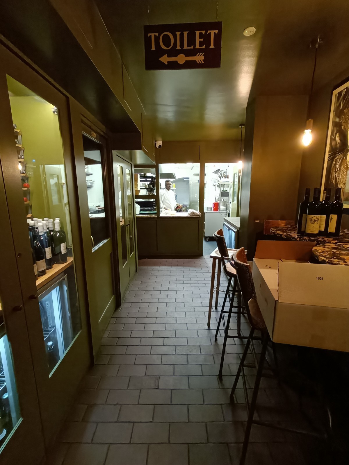 From bottom of stairs, looking straight. Wine display on left. Bathroom door behind. Open kitchen at back.