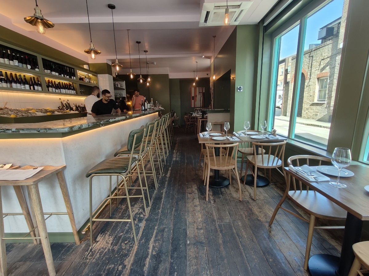 From entrance facing right. Tables and chairs along right wall. High table and stools behind. Open kitchen and stools along left.