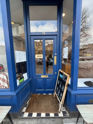 From pavement facing venue. One small step up to blue door with glass panel. Push on right. A-frame sign on top right of step.