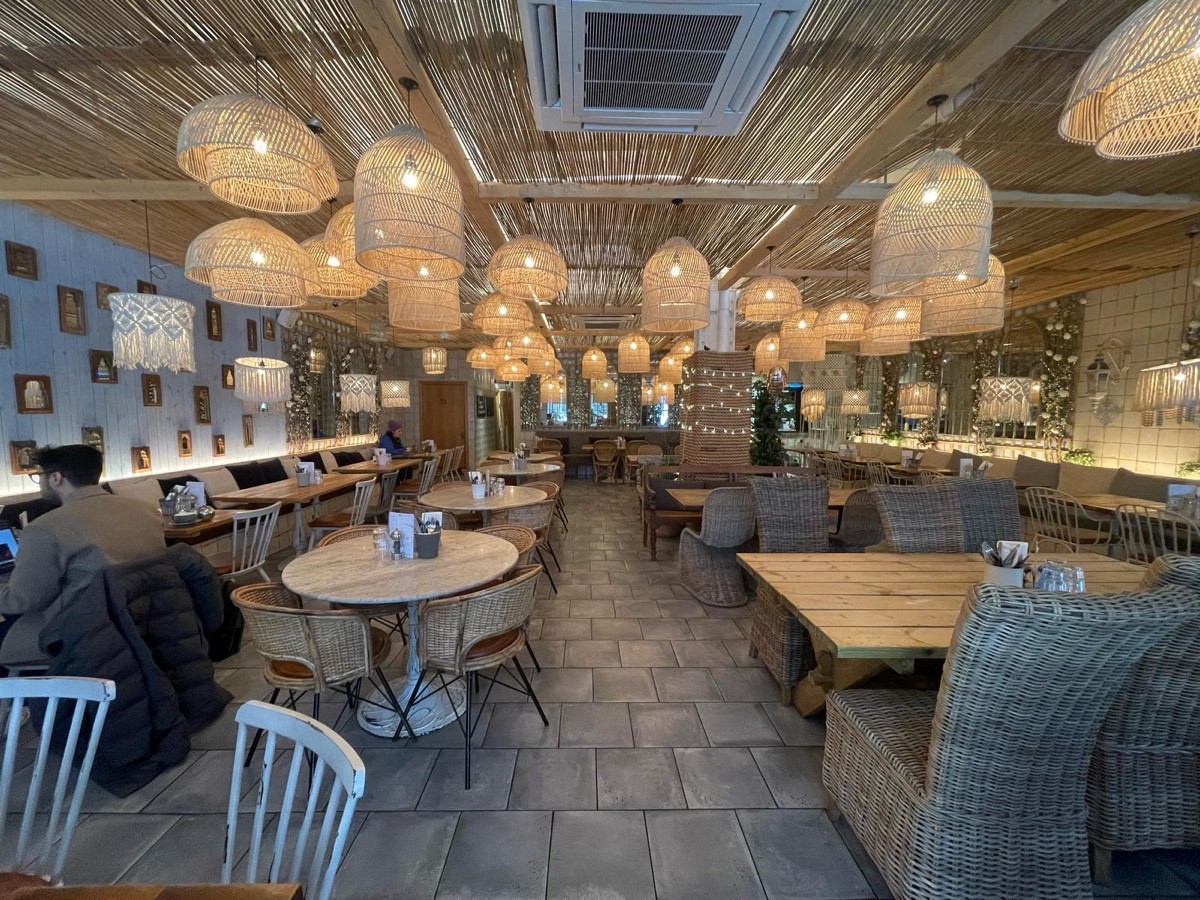 From opening facing straight. Half booth along left. Tables with chairs in a row to left and right. Half booth on right wall. Toilet on back left.
