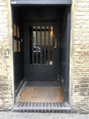 From pavement facing venue. Step up to opening ahead followed by doormat. Black door with glass panel at back.