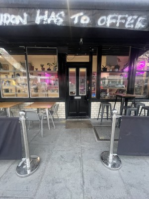 From street facing venue. Opening ahead. Restaurant barriers on left and right for outdoor seating. Entrance door at back.