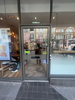 Glass door with black frame ahead. Push silver metal handle on right to open. Assistance call button on right of door.