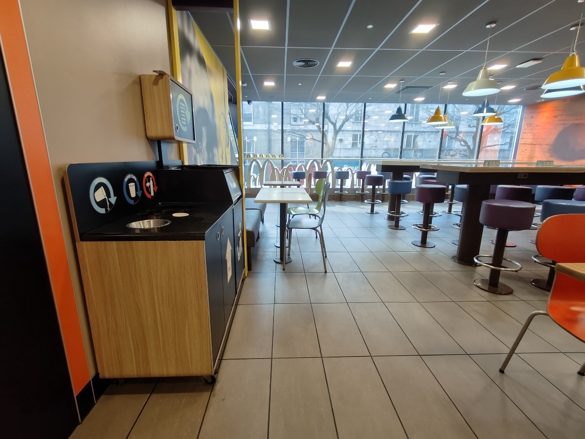 From doors facing straight. Bin on left. Half booth behind. Path ahead to more seating. Stools and high table at back right.