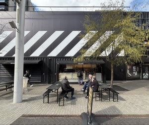 From street facing venue. Standing sign ahead. Path behind to counter on back. Outdoor seating along both sides.