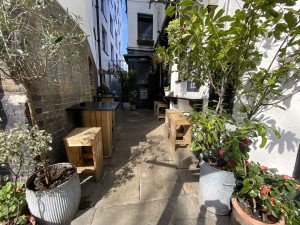 From street facing venue, opening ahead. Plants on left and right. High tables along left and right wall. Entrance on back right.