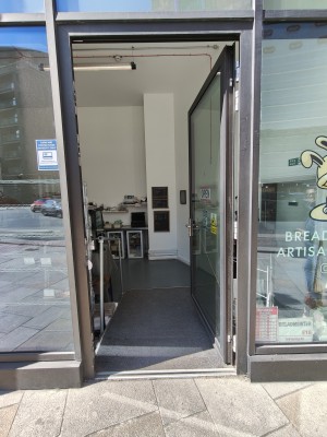 Grey framed glass door. Push silver handle on left to open. Ramp going down after door. Grey handrail on left.
