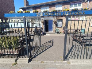 From street, facing venue. Black, metal gates ahead. Push from right to open. Outdoor seating after. Doorway at back.