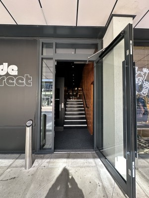 From pavement facing door to left of Side Store venue. Glass door with black frame ahead. Door opens outwards from left. Door push button to left.