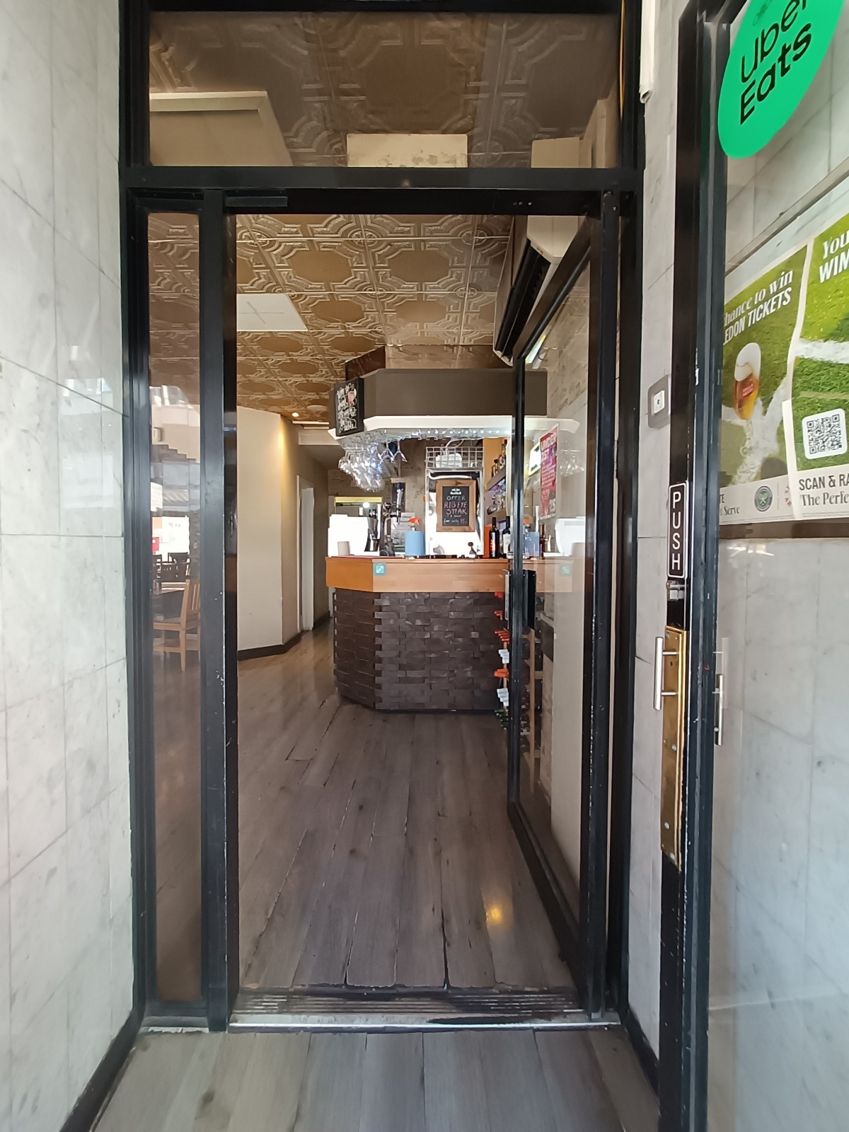 From top of step, looking straight. Glass door with black, metal frame ahead. Push from left to open.