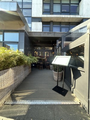 From street facing venue. Standing menu then planter on right. Entrance behind at back right. Seating at back. Planter on left.