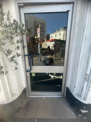 Glass door with metal frame and blue handle. Push from left to open.
