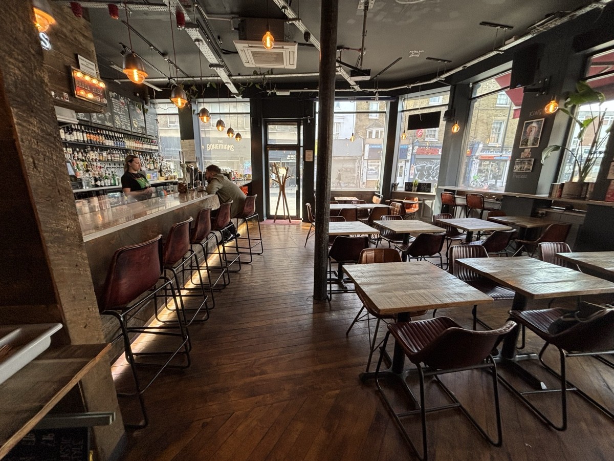 From start of bar counter facing right. The bar counter and stools along left. Tables and chairs along right. Pillar on back right.
