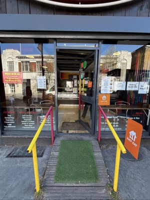 A ramp up with grass mat ahead to door. Yellow and red handrails on either side of it. Glass door with black frames on top. Push left handle to open.