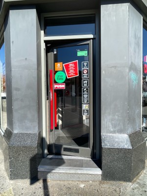 Facing glass door with gray frame. Push red handle on left.