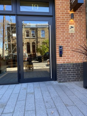 Glass door with black frame ahead. Call reception using call button on right hand wall then push from metal handle on right to open