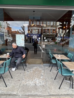 From sidewalk facing venue. Path ahead to glass entrance door on back. Push door to open via handle on right. Outdoor seating along both sides.