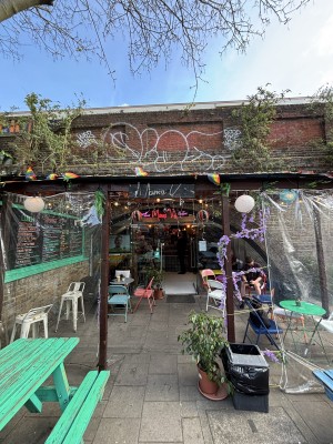 From pavement facing venue. Open doorway ahead. Outdoor seating along left and right.